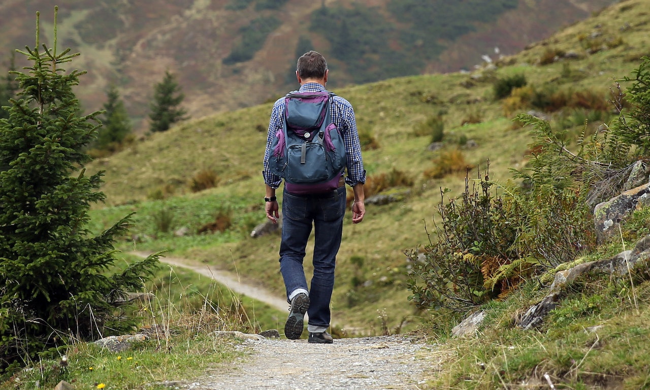 Man Carrying Backpack Walking Near Green Tree and Grass Duriong Daytime